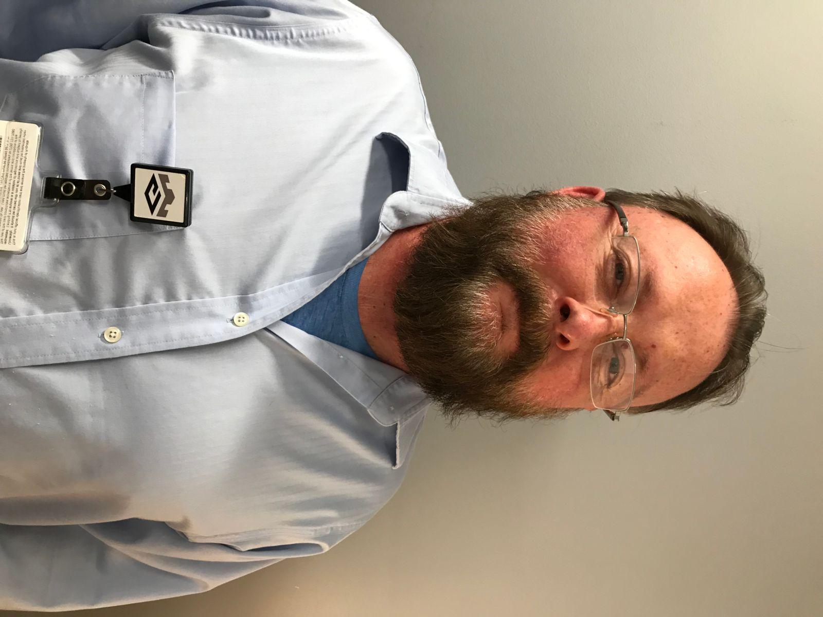 A Caucasian male with brown hair and a beard, Chester smith has rimless glasses and is wearing a blue tshirt under a light blue, collared, button down dress shirt.