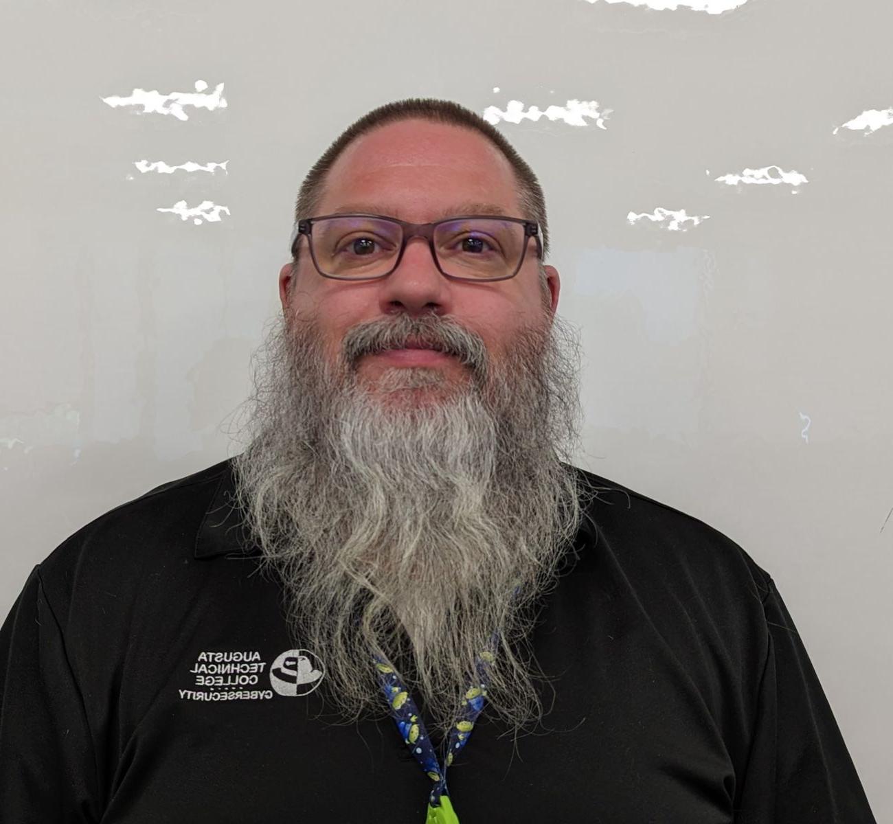 A Caucasian male with short brown hair and grey beard wearing a black Augusta Technical College Cybersecurity shirt. 