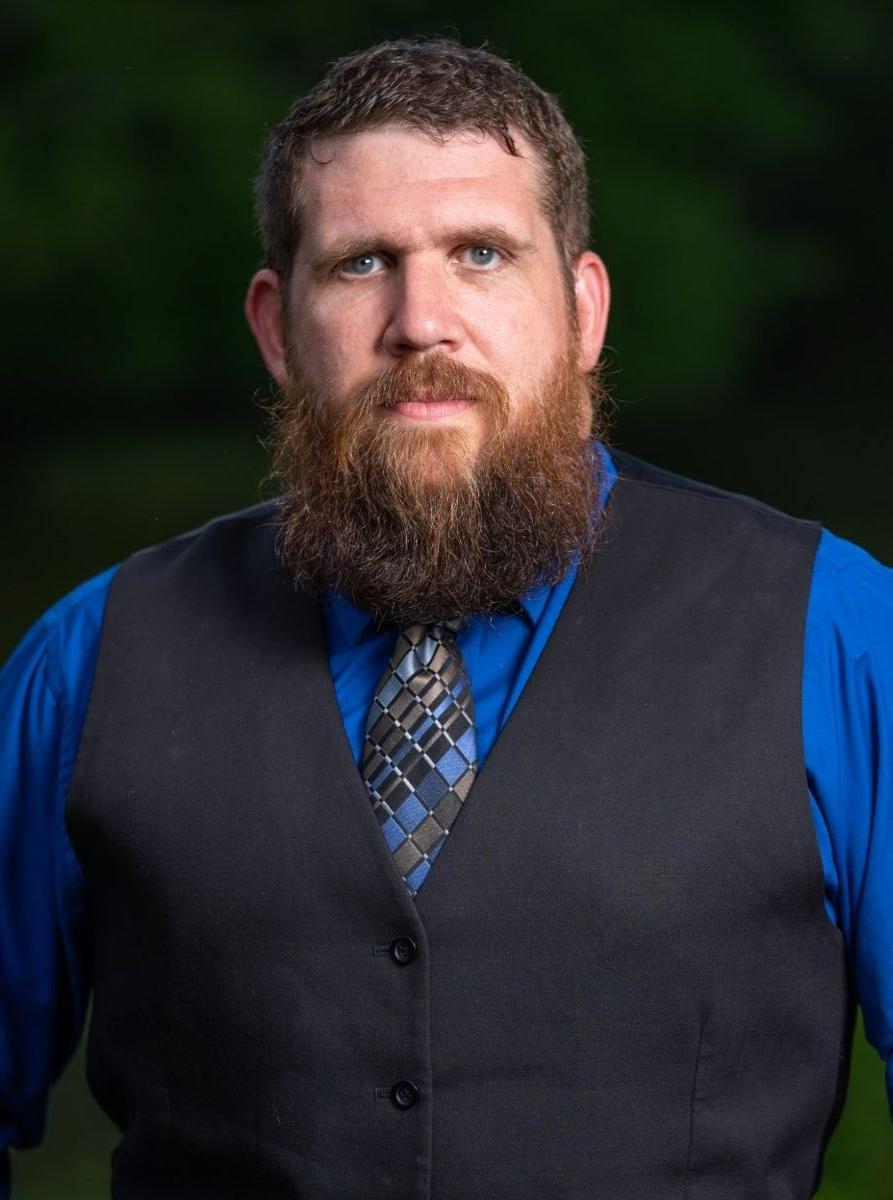 A Caucasian male with brown hair, brown beard, wearing a blue dress shirt and vest with tie with a light blue gold black and blue checkered pattern.