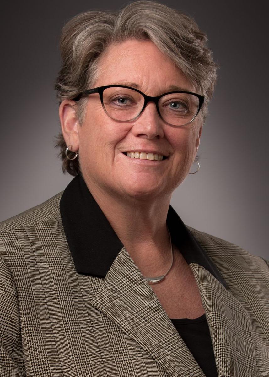 Dean Kellie McElroy Hooper, a Caucasian female with short grey and brown hair, smiles wearing glasses, a brown suit jacket with a black collar, and a black shirt.